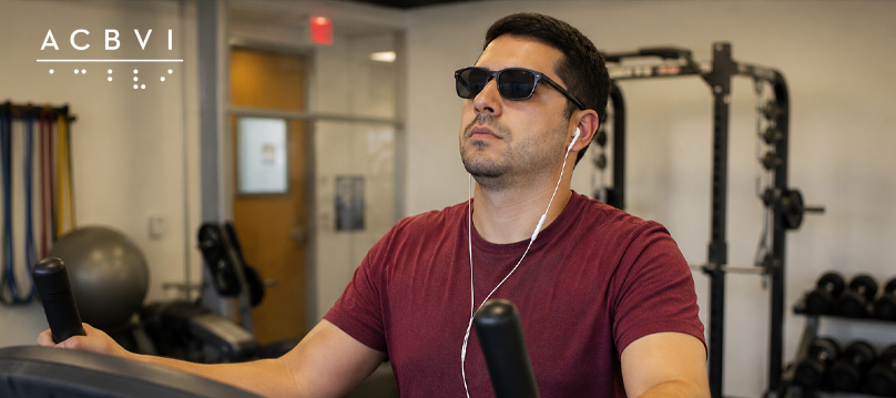 A visually impaired man wearing dark sunglasses and earbuds uses an elliptical machine in a gym, with weights and fitness equipment visible in the background.