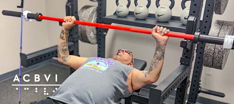 Client using adaptive gym equipment performs a bench press exercise during a fitness session at ACBVI, with tactile markers and strength-training equipment visible in the background.