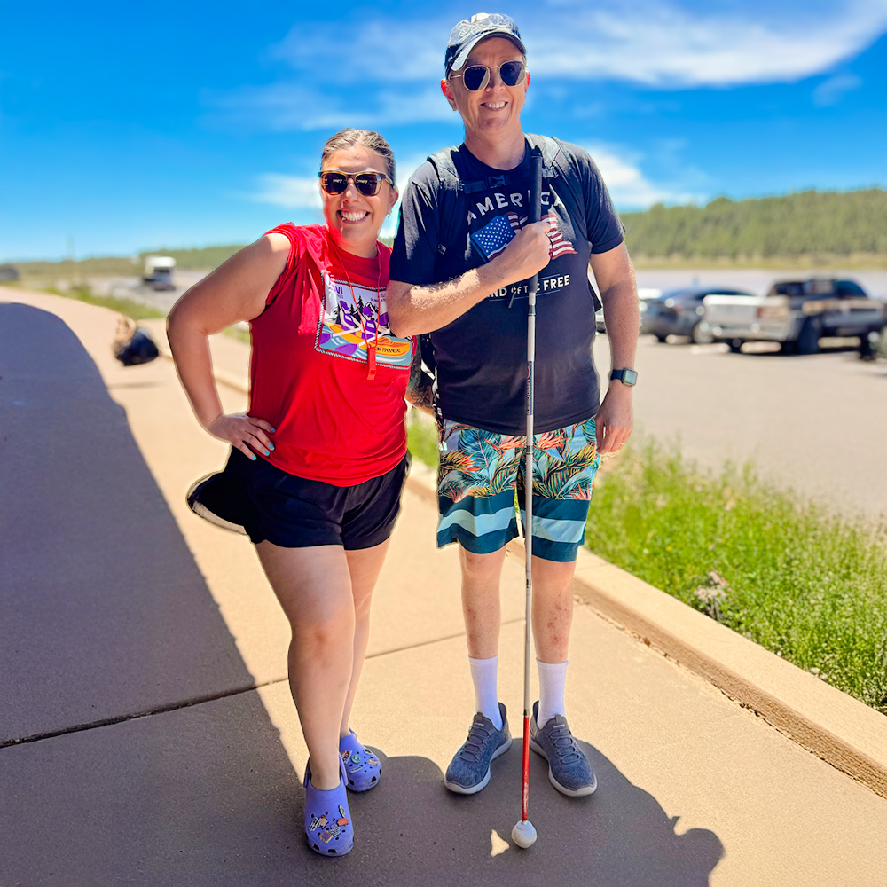 Man using a white cane walks with a smiling woman outdoors.